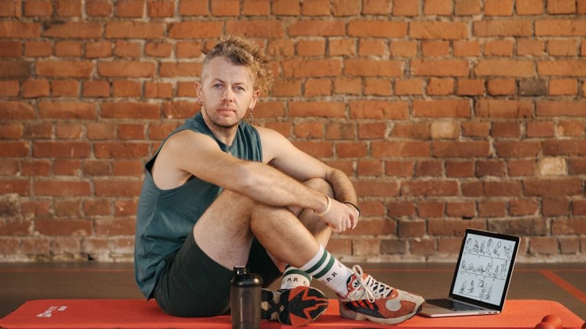 A man sits on an exercise mat with a laptop showing workout routines, suggesting a fitness regimen.